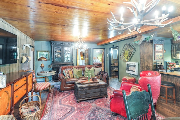 A cozy living room with a wooden ceiling, a large chandelier, a red leather sofa, a brown leather armchair, and a vintage suitcase coffee table.
