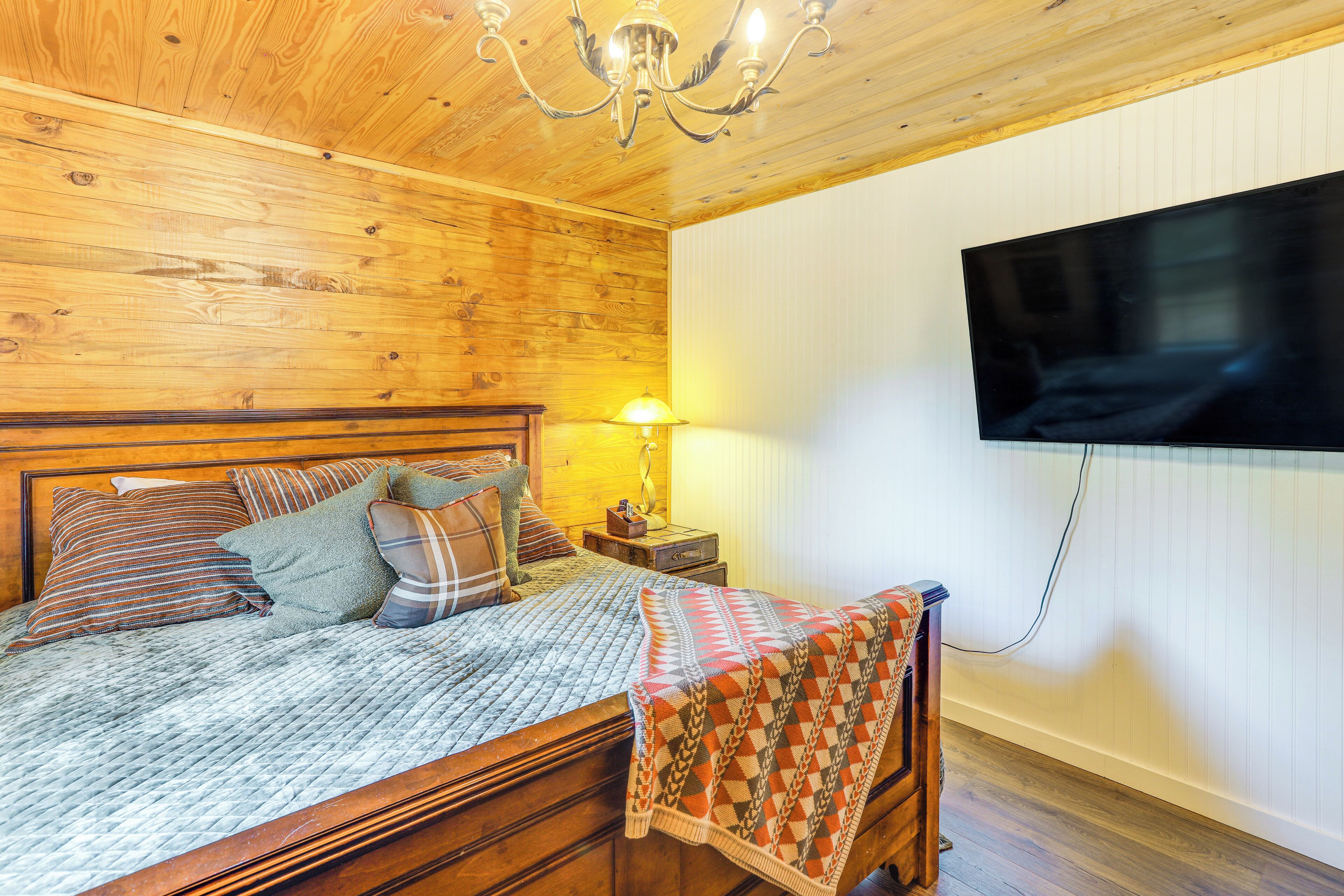 Quarto com painéis de madeira, uma cama, televisão fixada na parede e mesa de cabeceira com abajur.