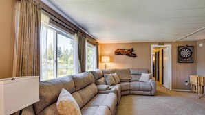 A modern living room with a sectional sofa, a lamp, a dartboard, and a view through large windows. - Trails End Retreat in Belfair w/ Private Dock (Belfair)