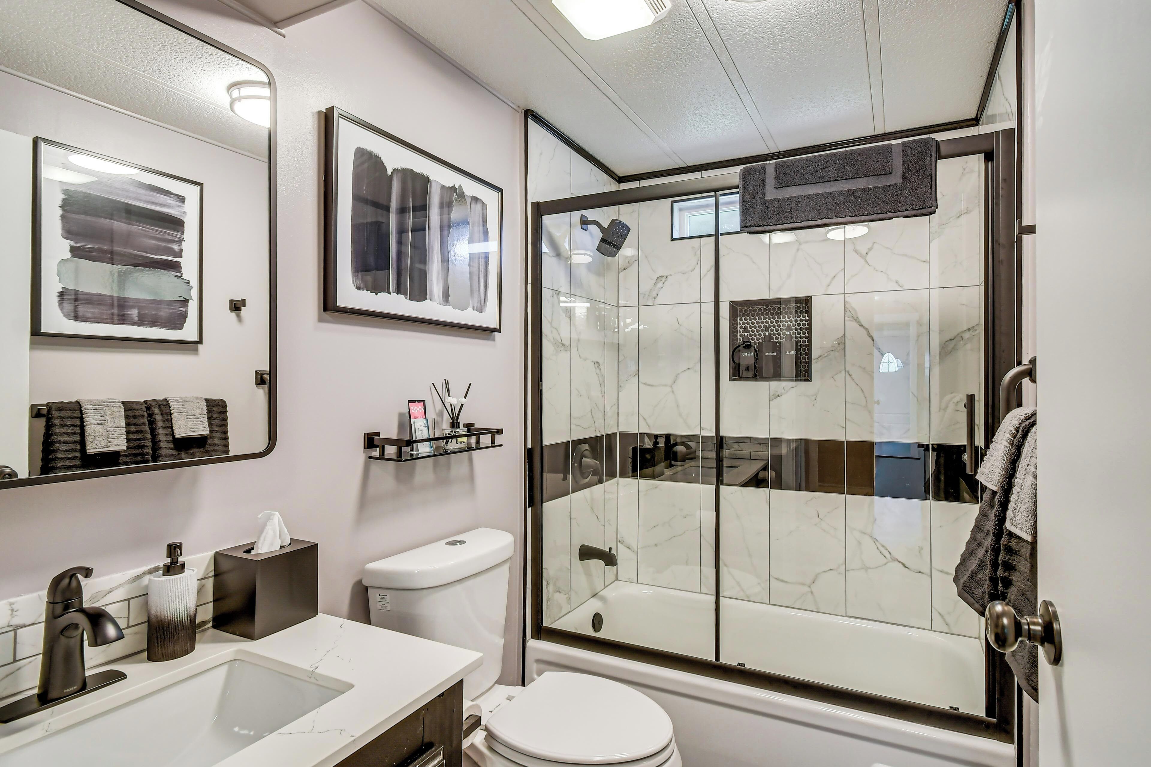 A modern bathroom with a glass-enclosed shower, a white toilet, a sink with a marble countertop, and framed artwork on the wall.