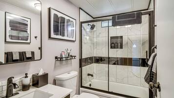 A modern bathroom with a glass-enclosed shower, a white toilet, a sink with a marble countertop, and framed artwork on the wall.