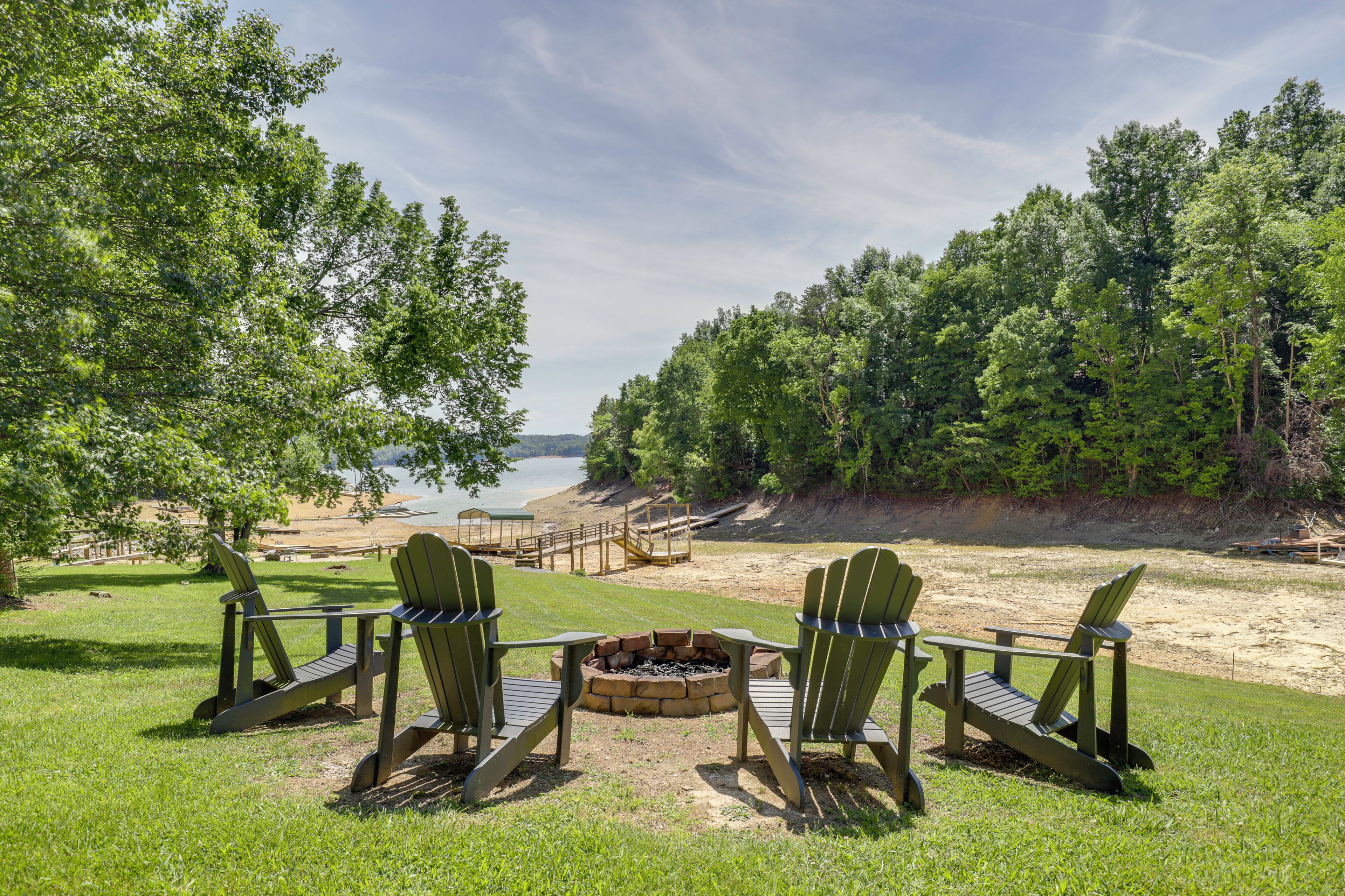 A lakeside view with three Adirondack chairs facing a fire pit, surrounded by lush greenery and a wooden deck.