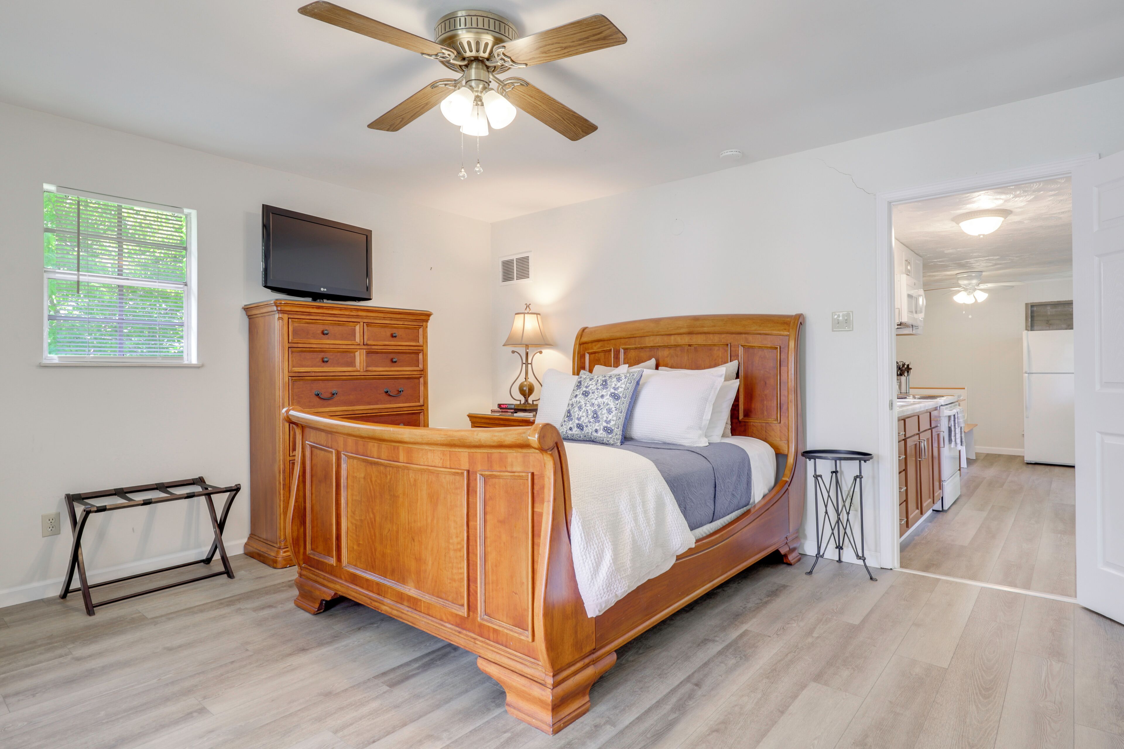 A bedroom with a wooden bed, a ceiling fan, a TV, a dresser, a lamp, and a view of the kitchen.