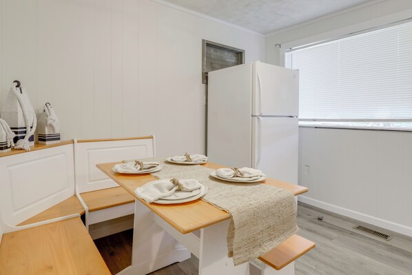 Una pequeña kitchenette con un refrigerador blanco, una mesa de madera dispuesta con platos y servilletas, y un banco.