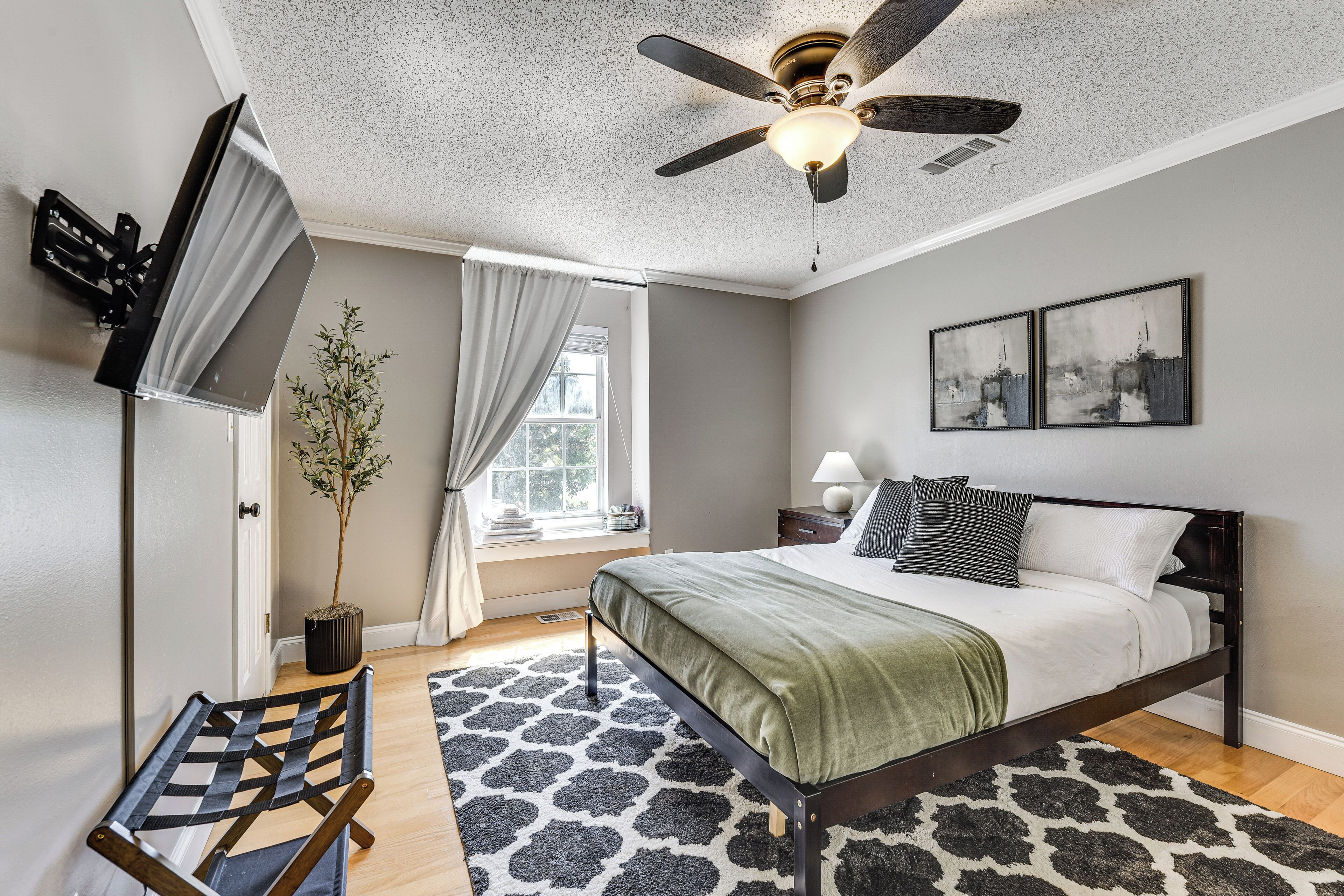 A bedroom with a bed, a ceiling fan, a window with curtains, a potted plant, and artwork on the wall.