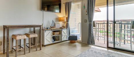 Ein Zimmer mit einer Schiebeglastür, die auf einen Balkon führt, einem Holztisch mit Hockern, einem Schrank mit Stauraum und einem an der Wand befestigten Fernseher.