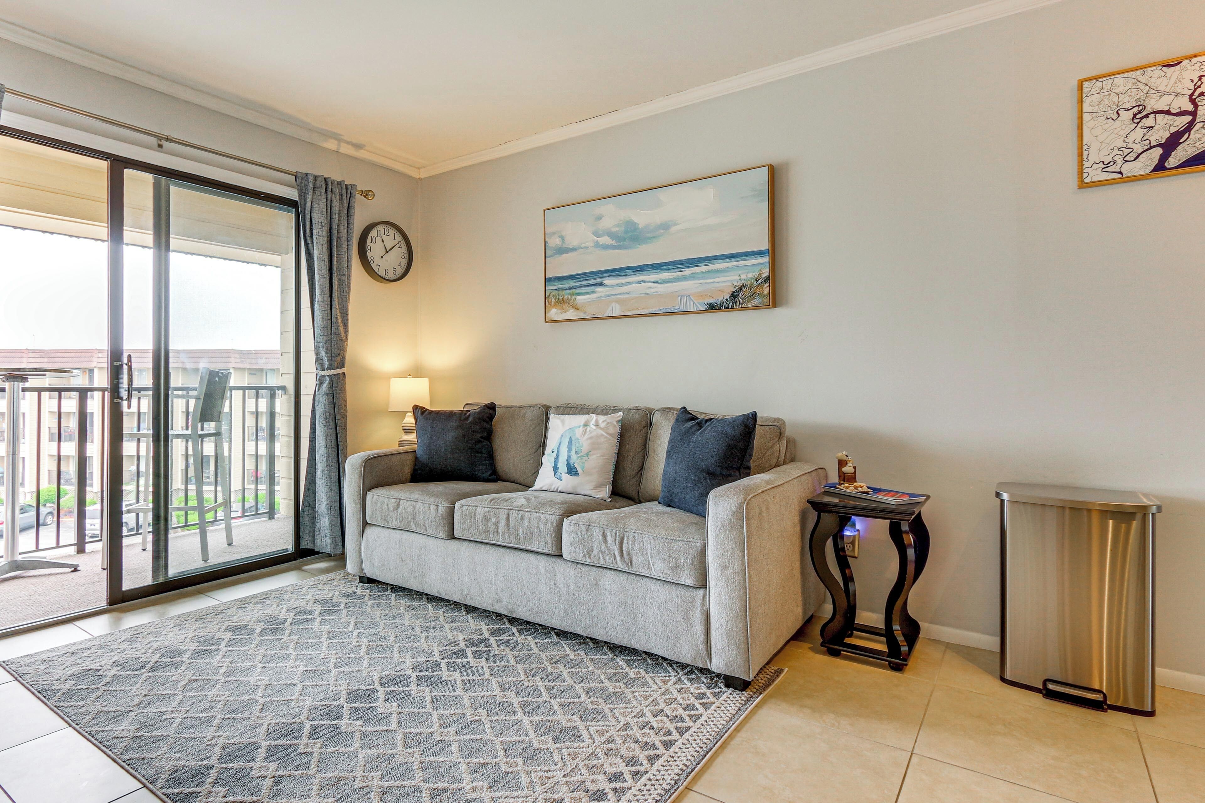 A living room with a sofa, a rug, a painting, a trash bin, and a view through a glass door.