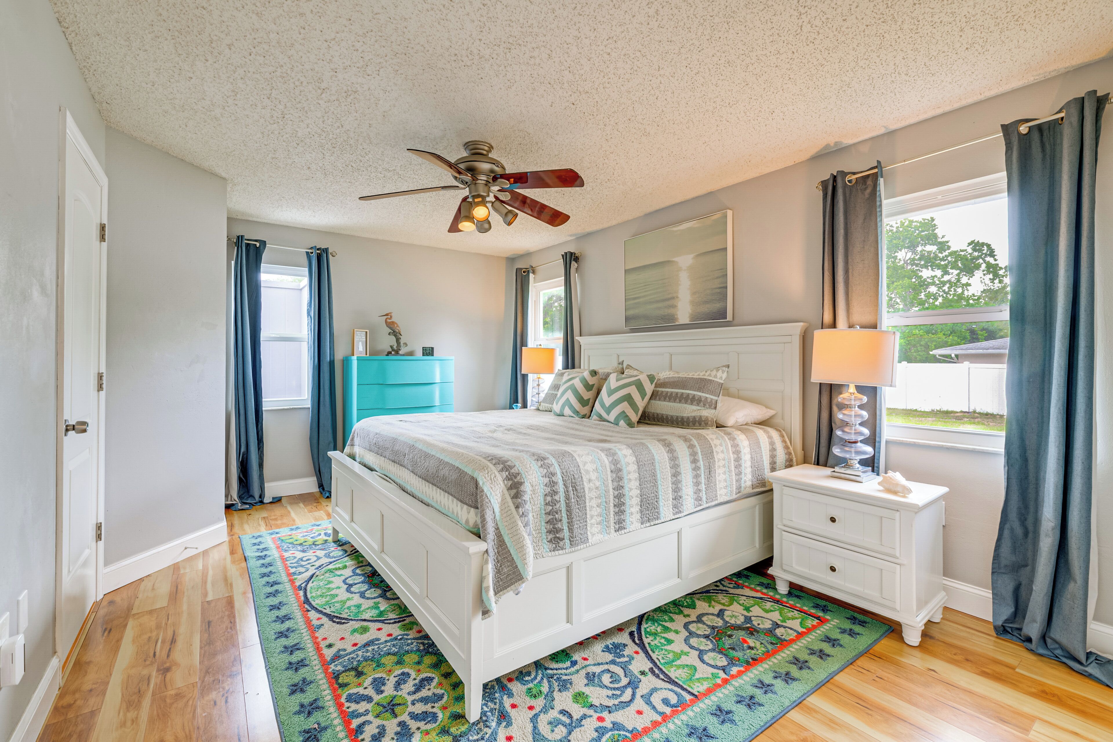 A bedroom with a bed, nightstand, ceiling fan, window with curtains, and a view of greenery outside.