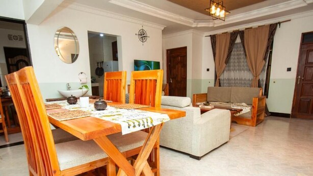 A dining area with a wooden table and chairs, a sofa, and a kitchen in the background. - Ukarimu Hosts (Arusha)