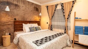 A bedroom with a wooden headboard, a patterned bedspread, and a window with patterned curtains. - Ukarimu Hosts (Arusha)
