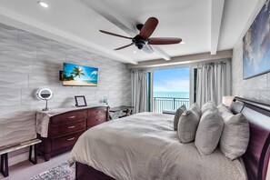 Ein Schlafzimmer mit Bett, Deckenventilator, Fernseher, Schminktisch mit Spiegel und Meerblick.