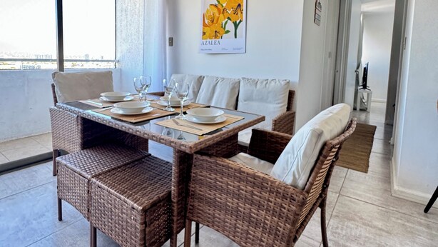 A dining area with wicker furniture, a table set for four, and a wall-mounted picture of yellow flowers. - Departamento 1501 Movistar Arena (Santiago)