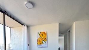 A dining area with a table set for four, wicker chairs, a ceiling light, and a wall-mounted picture of yellow flowers. - Departamento 1501 Movistar Arena (Santiago)