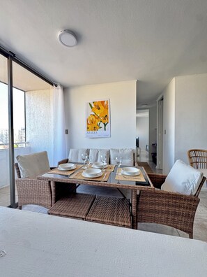 Un comedor con una mesa dispuesta para cuatro, sillas de mimbre, una lámpara en el techo y un cuadro colgado en la pared con flores amarillas.