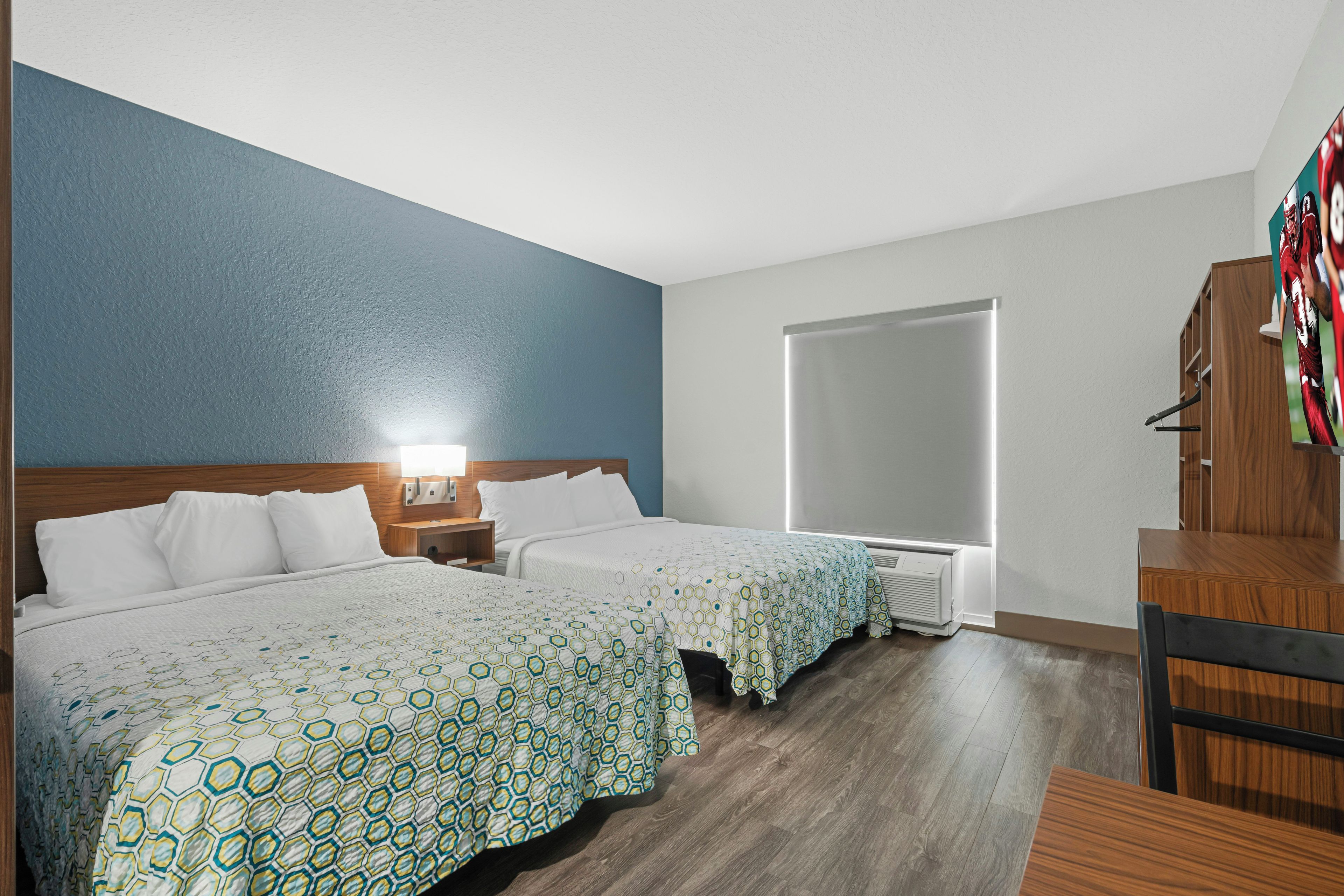 A hotel room with two beds, a desk, a chair, a TV, and a window.