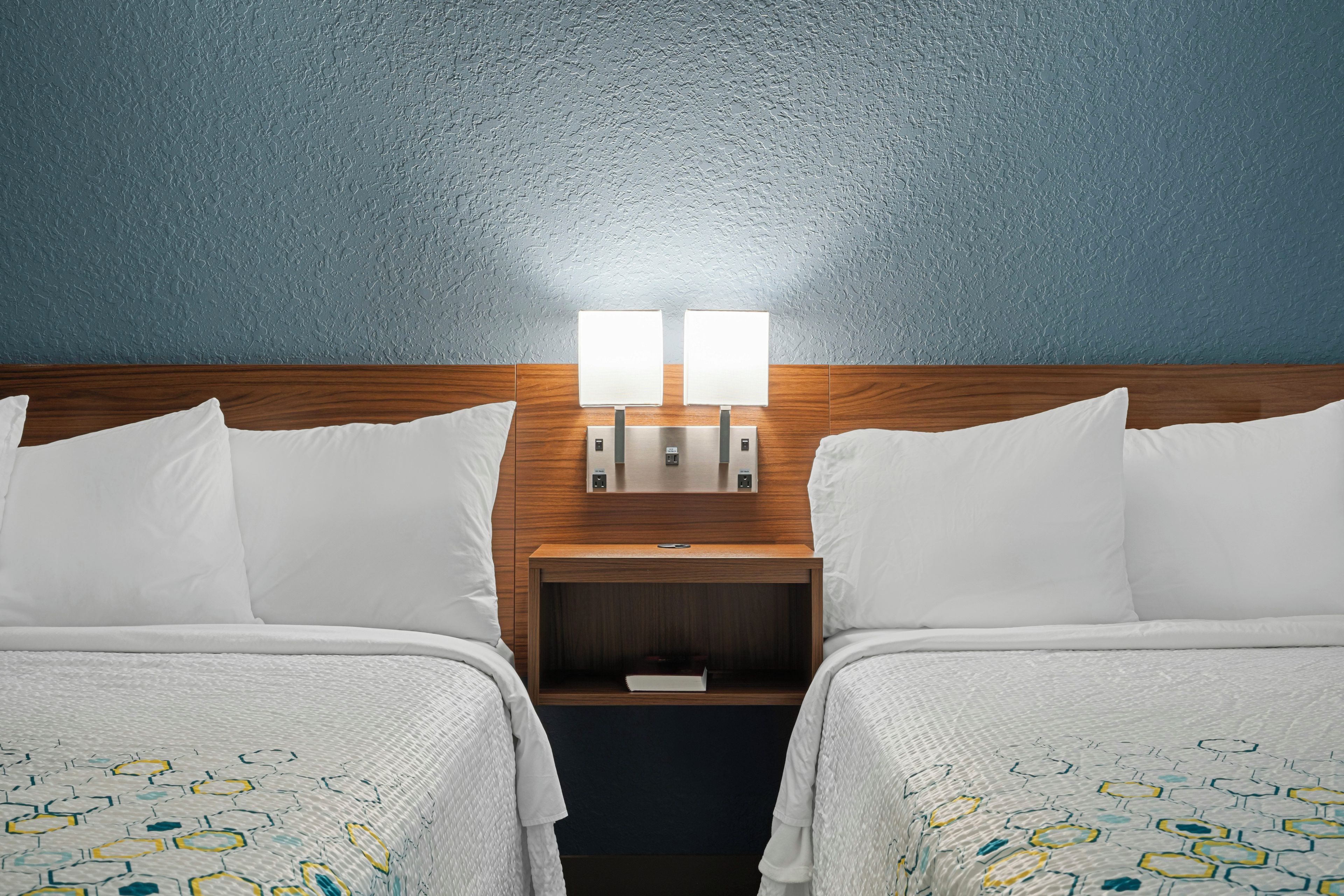 Quarto de hotel com duas camas, cabeceira de madeira, mesa de cabeceira com abajur e parede azul com textura.