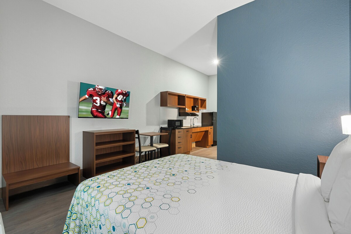 Una habitación de hotel con cama, escritorio, silla, kitchenette y televisor montado en la pared.