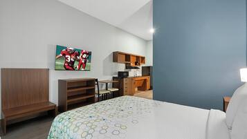A hotel room with a bed, a desk, a chair, a kitchenette, and a wall-mounted TV.
