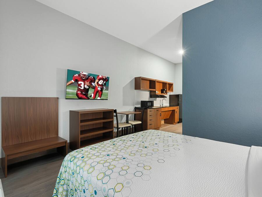 A hotel room with a bed, a desk, a chair, a kitchenette, and a wall-mounted TV.