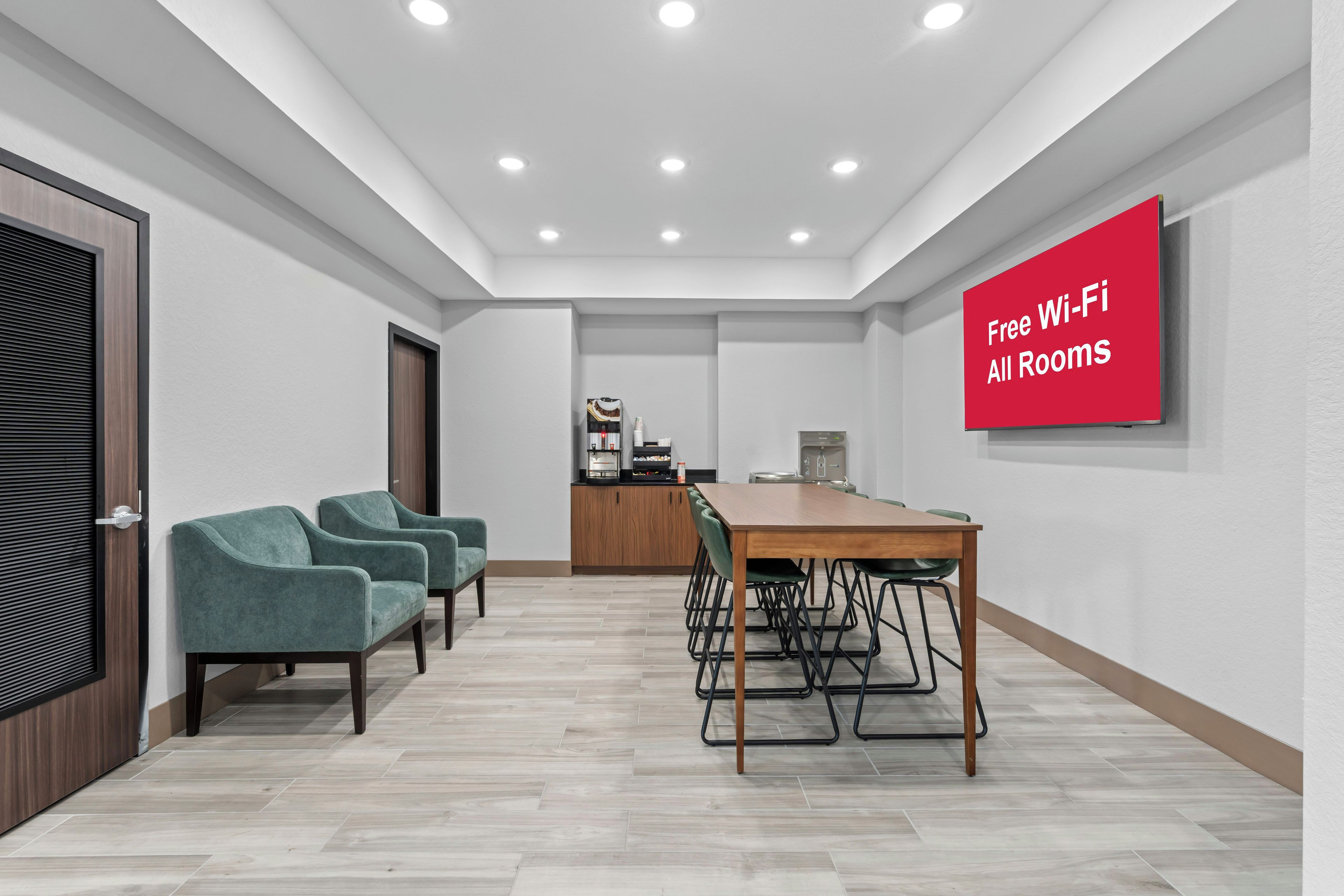 A conference room with a wooden table and chairs, a red sign stating 'Free Wi-Fi All Rooms,' and a kitchenette in the background.