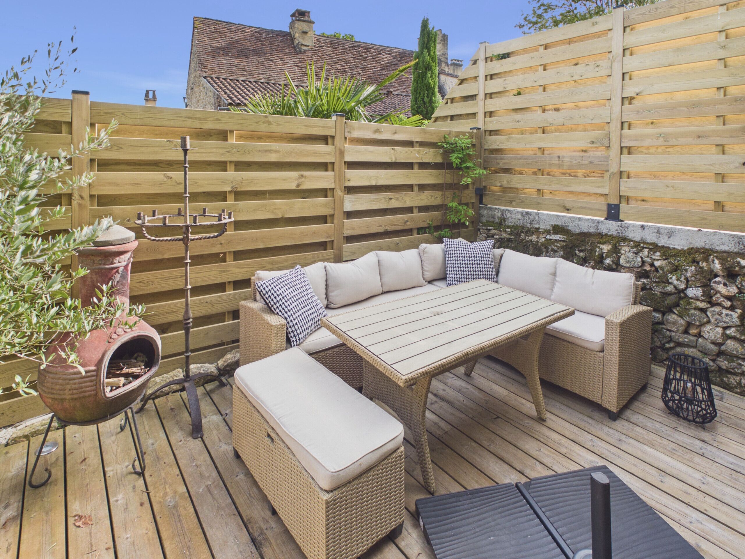 An outdoor patio with a wooden table and wicker furniture, surrounded by a wooden fence and stone wall.