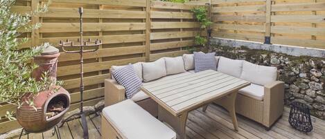 An outdoor patio with a wooden table and wicker furniture, surrounded by a wooden fence and stone wall.