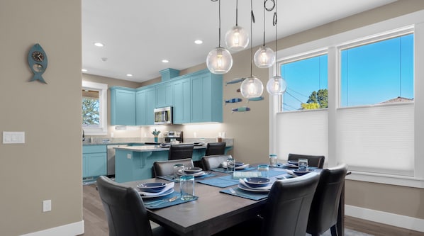 A modern kitchen with teal cabinets, a dining area with a table set for four, and a large window with blinds. - Mac's Surf Shack at Seaside Beach (Seaside)