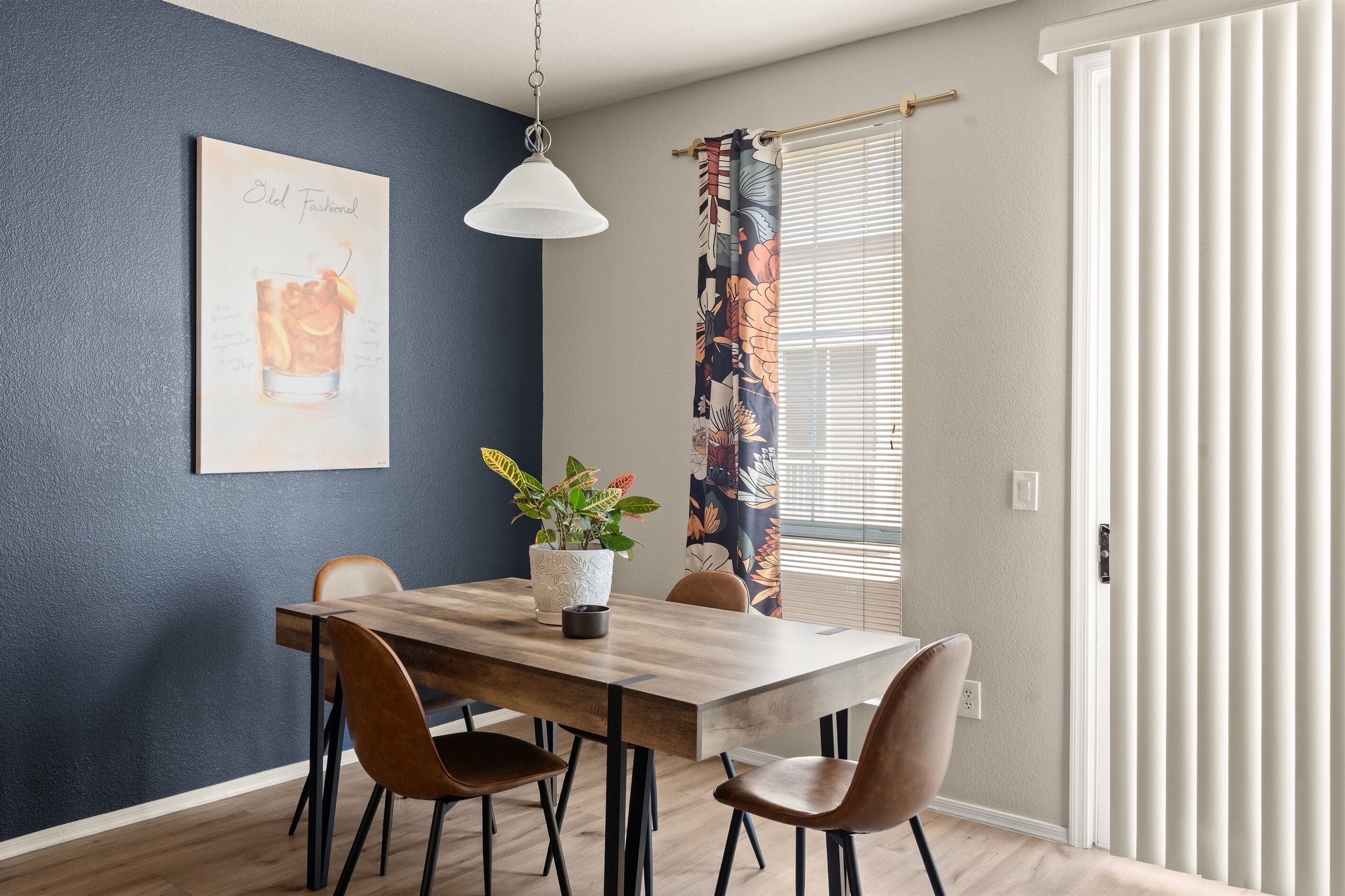 Un coin repas comprenant une table en bois, quatre chaises, un luminaire suspendu, une plante en pot et une fenêtre avec des stores.
