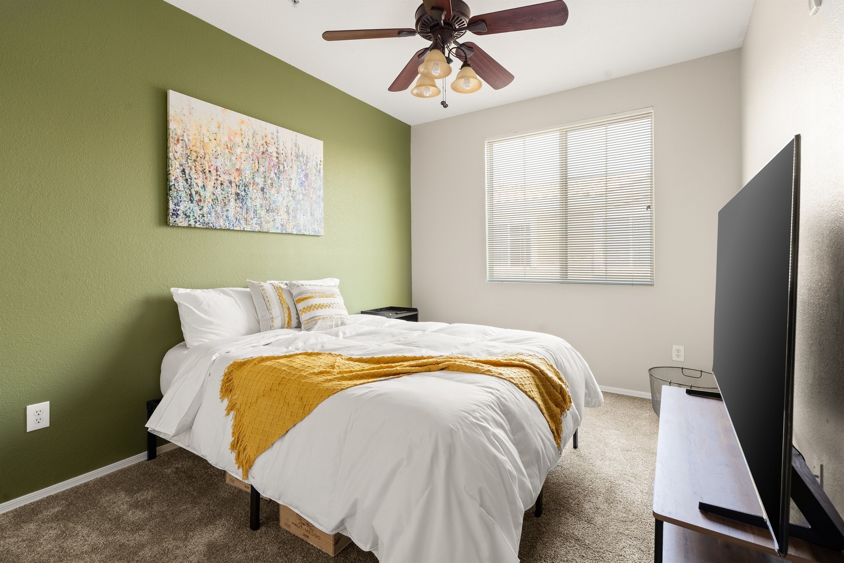 A bedroom with a bed, a ceiling fan, a window with blinds, a painting on the wall, and a TV on a stand.