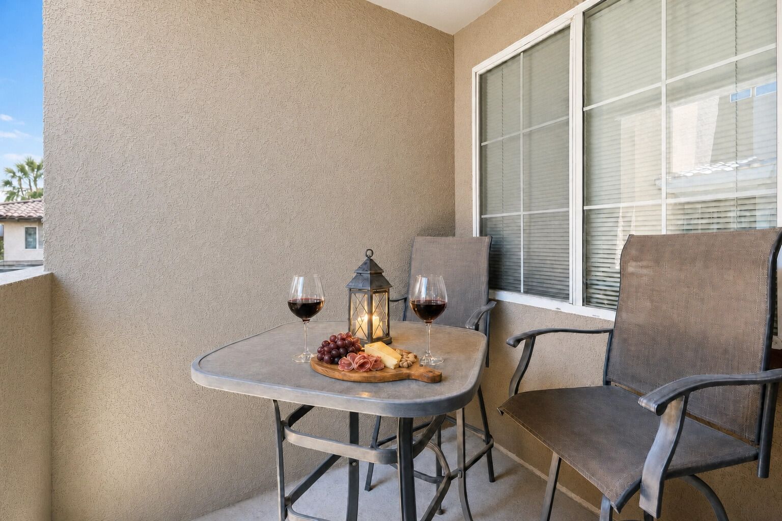 A balcony with a table set for two, featuring a candle, two glasses of red wine, and a plate with grapes and cheese.
