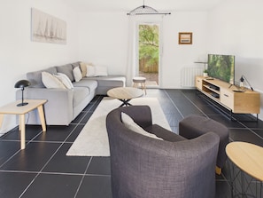 Ein modernes Wohnzimmer mit grauem Sofa, einem runden Couchtisch, einem grauen Sessel, einem Fernseher auf einem Holzgestell und einer Tür, die nach draußen führt.