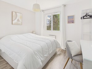 Ein Schlafzimmer mit Bett, Schreibtisch, Stuhl, Kleiderhaken und einem Fenster mit Blick auf Bäume.