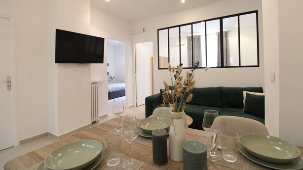 A dining area with a table set for four, a green sofa, a flat-screen TV mounted on the wall, and a large window. - Calm Elegant Apartment in the Heart of Nice (Nice)