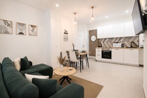 Ein modernes Wohnzimmer mit einem grünen Sofa, einem Essbereich mit Stühlen, einer Küche mit weißen Schränken und einem an der Wand montierten Fernseher.