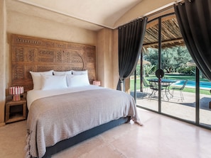 Un dormitorio con una cama grande, cabecera de madera, mesita de noche con lámpara y vista a una piscina exterior y mobiliario de patio.