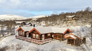 A wooden cabin with a porch and a garage, surrounded by snow-covered hills and trees.