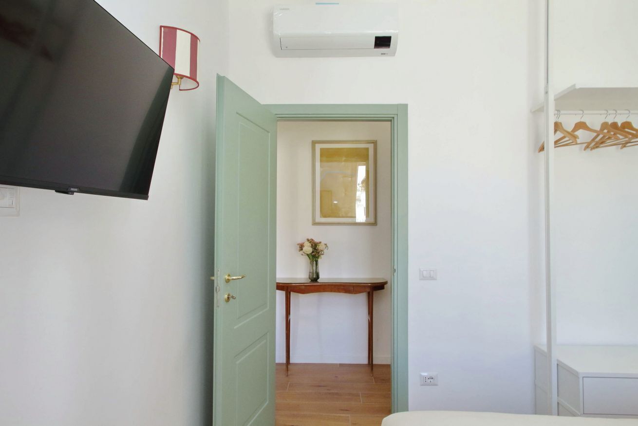 A room with a bed, a TV, a wall-mounted air conditioner, a door leading to another room, and a small table with a vase of flowers.