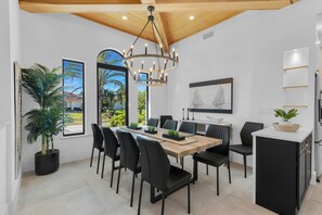 A modern dining area with a wooden table, black chairs, a chandelier, and a view of the outdoors. - Island Time on Ivory I 4 Bed Waterfront Home (Marco Island)