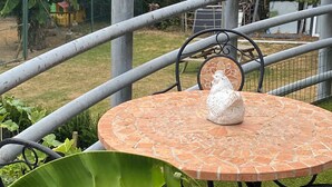 Una mesa redonda al aire libre con un objeto blanco encima, rodeada de sillas de metal, con vistas a un jardín y una valla.