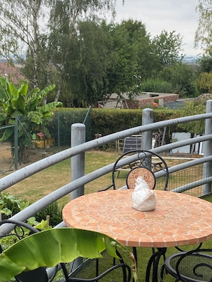 A round outdoor table with a white object on it, surrounded by metal chairs, with a view of a garden and a fence.