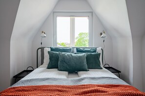 Ein Schlafzimmer mit schrägen Wänden, einem großen Fenster, einem Bett mit vielen Kissen, zwei Nachttischen und zwei Wandlampen.