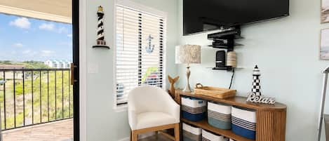 A room with a TV, a chair, a console table with baskets, a window with blinds, and a view of the outdoors.