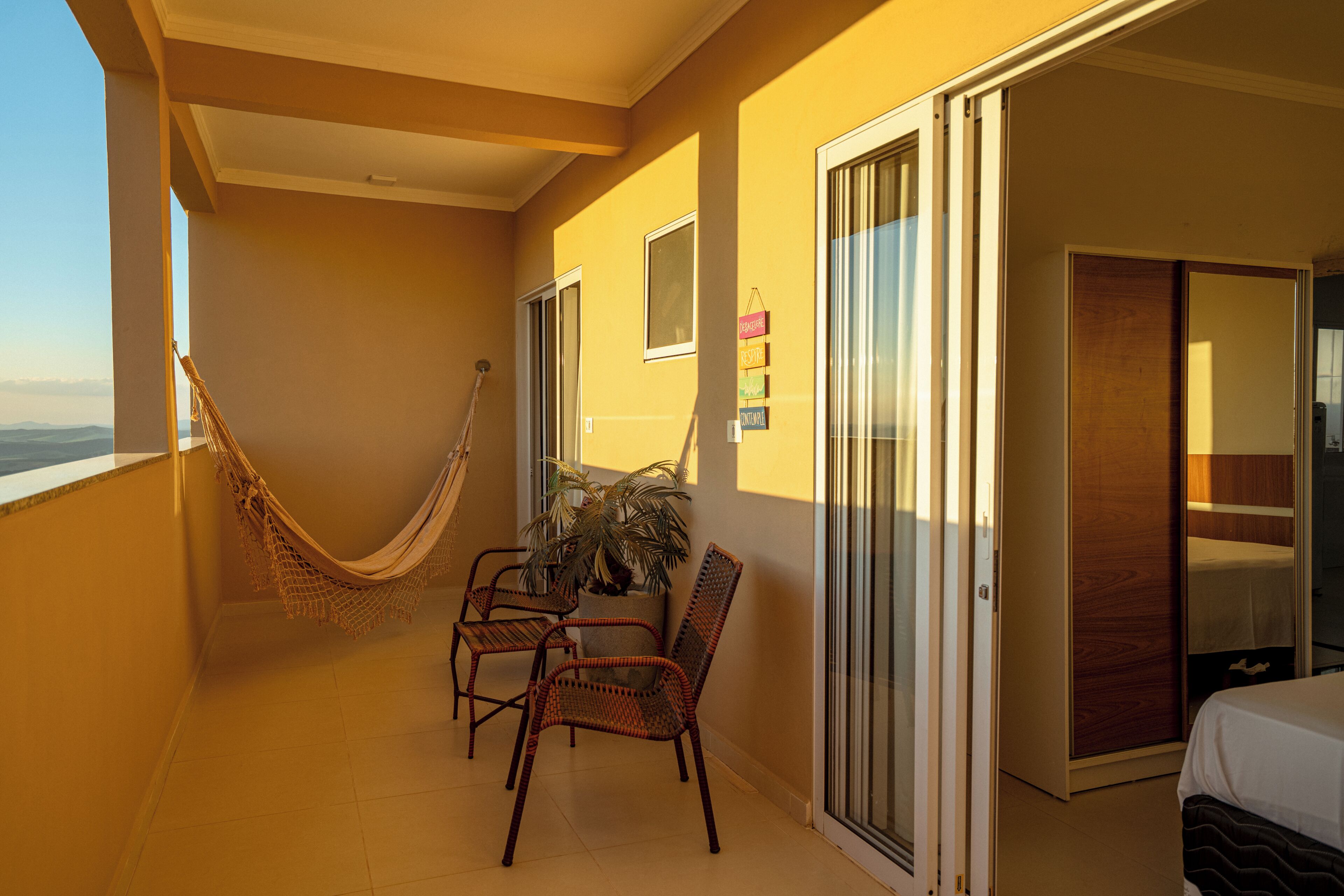 A balcony with a hammock, wicker chairs, a potted plant, and a sliding glass door leading to a bedroom.