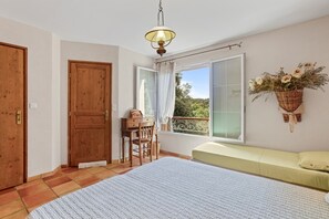 Ein Schlafzimmer mit Bett, Schreibtisch, Stuhl, einem Fenster mit Vorhängen, einem Balkon und einer Hängetasche mit Blumen.
