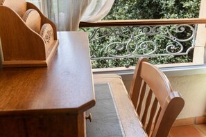 Une table en bois avec une chaise, une fenêtre ornée d’une rambarde en métal et une vue sur la verdure à l’extérieur.