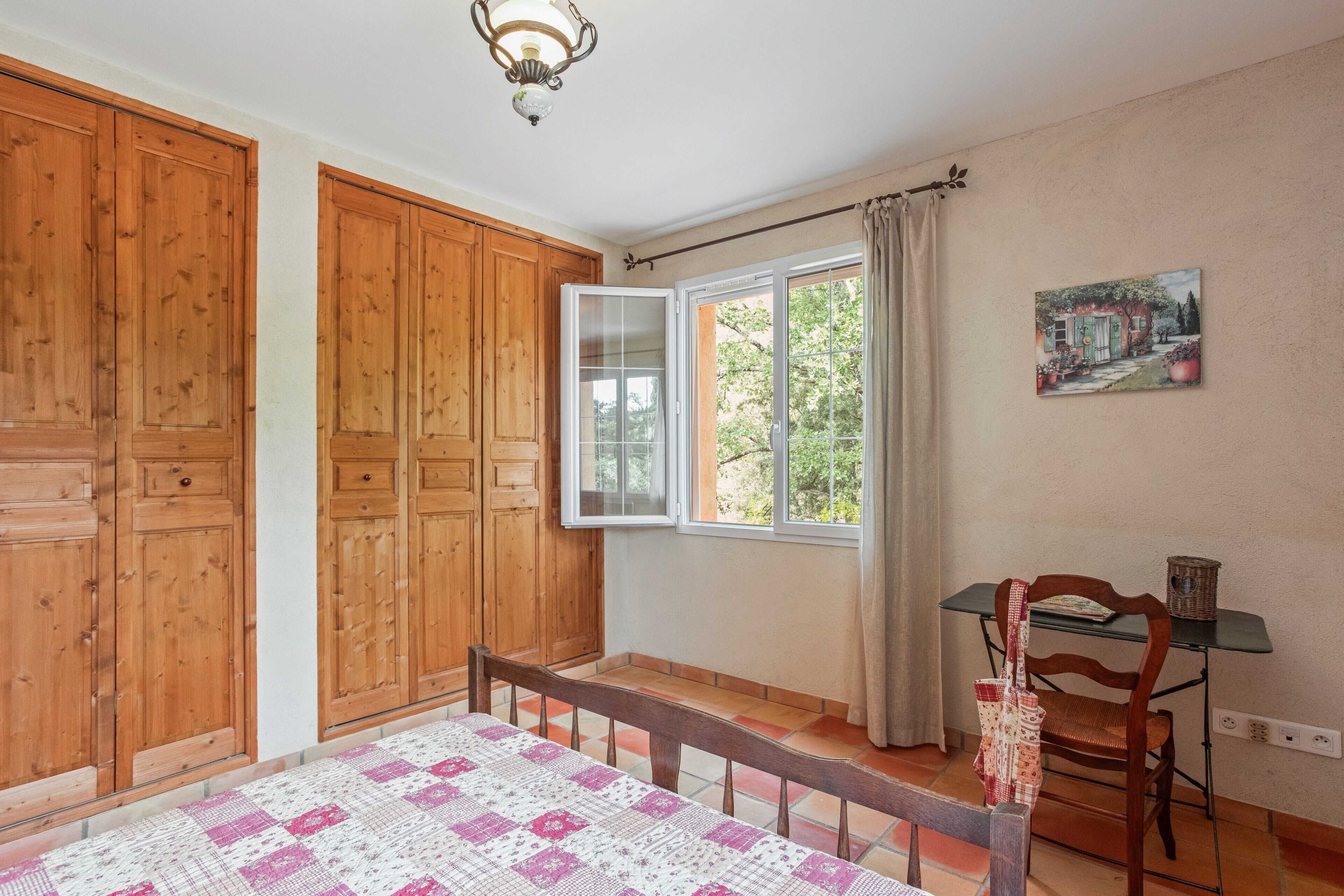 A bedroom with wooden cabinets, a bed with a quilt, a window with curtains, a table with a chair, and a wall-mounted lamp.