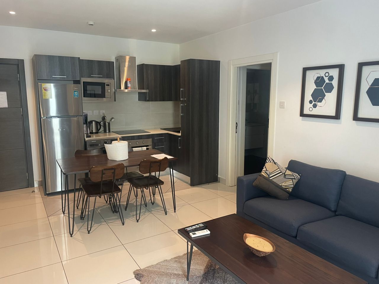 Uma cozinha moderna com geladeira, micro-ondas e fogão, uma área de refeições com mesa e cadeiras, e uma sala de estar com sofá e mesa de centro.