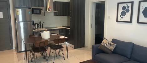 Uma cozinha moderna com geladeira, micro-ondas e fogão, uma área de refeições com mesa e cadeiras, e uma sala de estar com sofá e mesa de centro.
