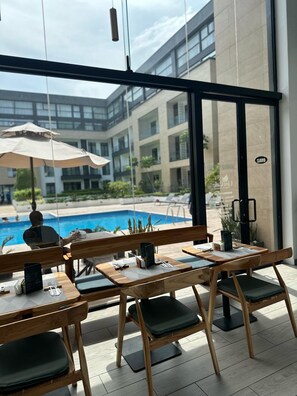 Un restaurante con mesas y sillas de madera, vistas a una piscina y un edificio moderno al fondo.