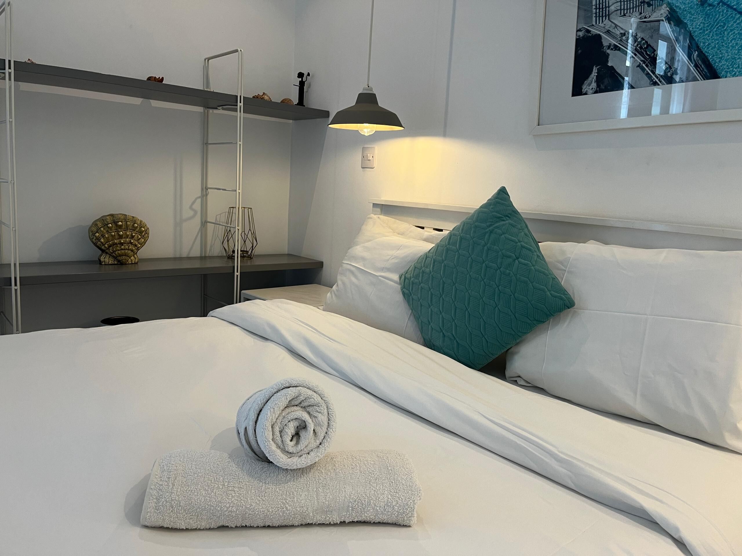 A neatly made bed with a rolled towel, a teal pillow, and a lamp on a bedside table.